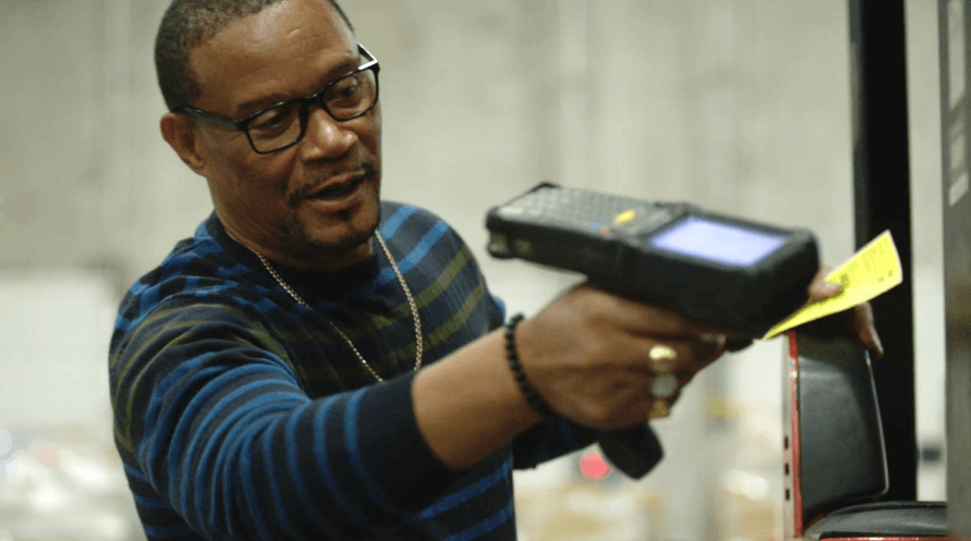 A man holding a scanner in a warehouse, surrounded by shelves and boxes.