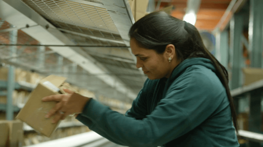 A woman in a warehouse picking up a box, surrounded by shelves filled with various items.